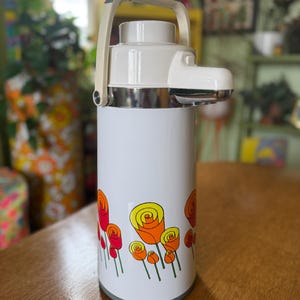 May include: A vintage white and chrome thermos with a cream-colored handle. The thermos features a floral design of red, orange, and yellow roses. The thermos is cylindrical and has a gray base.