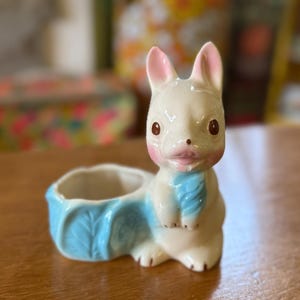 May include: A vintage ceramic bunny planter in cream with pink accents on the ears and cheeks. The bunny has brown eyes and a blue scarf. The planter section is a light blue color, shaped like a leaf.