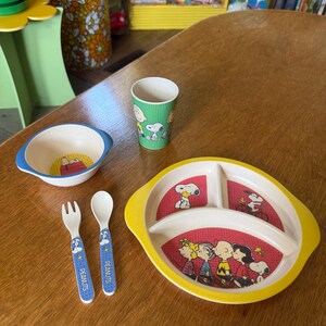 May include: A Peanuts-themed children's dining set including a yellow-rimmed divided plate, a blue-rimmed bowl, a green tumbler, and a matching fork and spoon. The items feature illustrations of Snoopy and the Peanuts gang on red backgrounds.