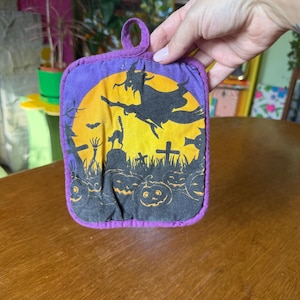 Peut inclure: Un dessous de plat carré en tissu avec une bordure violette et une boucle de suspension. Le centre présente un imprimé sur le thème d'Halloween avec la silhouette noire d'une sorcière volant sur un balai devant une grande lune jaune.