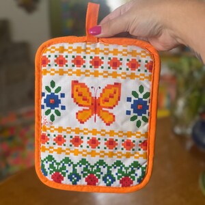 May include: A rectangular fabric pot holder with an orange border and hanging loop. The white surface features a pixelated orange butterfly, blue and green floral motifs, and rows of red and orange geometric patterns with a small C&C 79 logo.