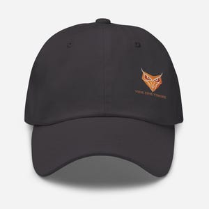 Peut inclure: Casquette de baseball gris foncé avec un logo de hibou brodé en orange et blanc. Le texte "VIDE SINE TIMORE" est en dessous. La casquette a une visière incurvée et un bouton sur le dessus.
