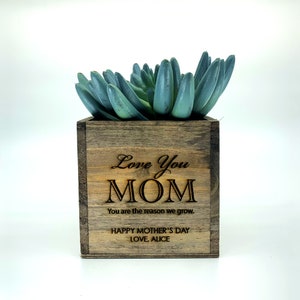 May include: A wooden planter with a succulent plant inside. The planter is engraved with the text "Love You MOM You are the reason we grow. HAPPY MOTHER'S DAY LOVE, ALICE".