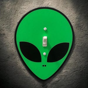 May include: A green alien-themed light switch cover with black eyes and a black outline. The switch is white and in the on position. The cover is mounted on a textured gray wall.