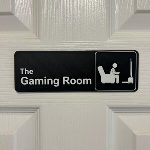May include: A black rectangular sign with white text that reads "The Gaming Room." A small graphic depicts a person seated in a chair, facing a television. The sign is mounted on a white door.