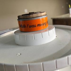 May include: A round orange container with a dark brown lid sits on a white tiered stand. The side of the container features black cursive text that reads "While I was musing the". The container is positioned centrally on the white plastic base.