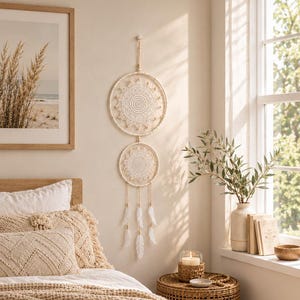 May include: A double-hoop wall hanging featuring intricate white crochet mandalas, suspended by a cord, with dangling white feathers attached to the lower hoop. The piece is crafted from off-white thread and features a symmetrical circular design.
