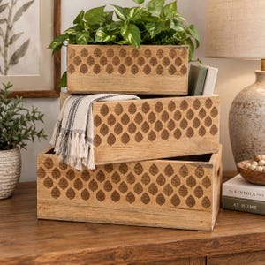 May include: A set of three rectangular light brown wooden storage crates stacked on top of each other. Each crate features a repeating dark brown leaf pattern etched across the front panel. A white and black striped fabric with fringe is draped.