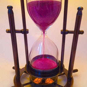 May include: An hourglass with a dark brown frame and a clear glass chamber filled with pink sand. The hourglass is supported by a decorative base with a compass design. The sand is flowing from the top chamber to the bottom.