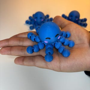 May include: A blue octopus figurine with a smiling face and rounded tentacles is held in a hand. Two other similar figurines are slightly out of focus in the background. The octopus has pink cheeks and purple accents.