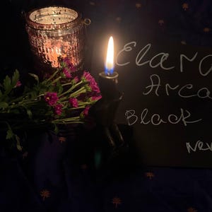 May include: A lit candle in a black holder with the text "Elara Arca Black" on a black surface. A pink glass jar with a lit candle and a bouquet of pink flowers are also in the image. The background is a dark blue fabric.