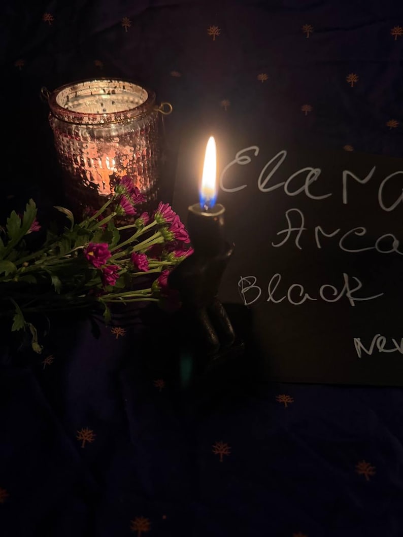May include: A lit candle in a black holder with the text "Elara Arca Black" on a black surface. A pink glass jar with a lit candle and a bouquet of pink flowers are also in the image. The background is a dark blue fabric.