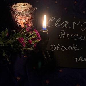May include: A lit candle in a black holder with the text "Elara Arca Black" on a black surface. A pink glass jar with a lit candle and a bouquet of pink flowers are also in the image. The background is a dark blue fabric.