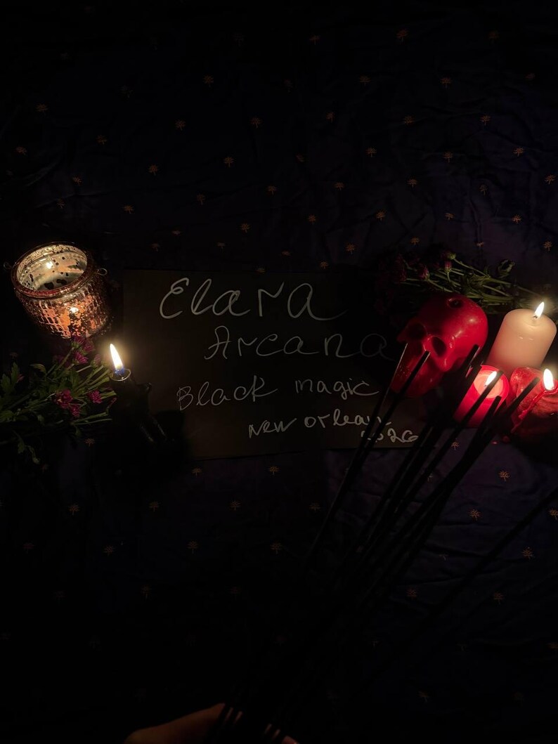 May include: A dimly lit scene featuring a chalkboard with the words "Elara Arcana Black Magic New Orleans". Lit candles, a red skull, and a decorative candle holder are visible. The background is a dark fabric with a subtle pattern.