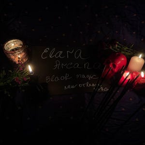 May include: A dimly lit scene featuring a chalkboard with the words "Elara Arcana Black Magic New Orleans". Lit candles, a red skull, and a decorative candle holder are visible. The background is a dark fabric with a subtle pattern.