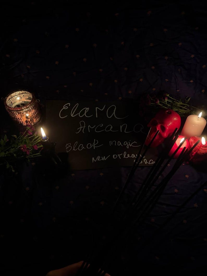 May include: A dimly lit scene featuring a chalkboard with the words "Elara Arcana Black Magic New Orleans". Lit candles, a red skull, and a decorative candle holder are visible. The background is a dark fabric with a subtle pattern.