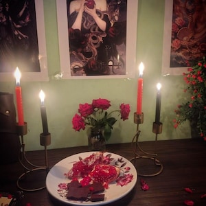 a table with a plate of food, a vase of roses, and three lit candles. The table is set against a green wall with framed artwork.