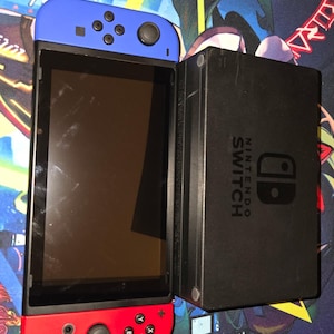 May include: A Nintendo Switch console with a black screen, blue left controller, and red right controller. The black dock is to the right. The console is on a colorful gaming mousepad.