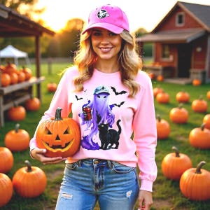 May include: A light pink long-sleeve sweatshirt featuring a graphic of a purple ghost wearing a cap, a black cat, and bats, paired with blue jeans and a pink baseball cap, while holding a carved orange jack-o'-lantern pumpkin.