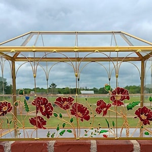 May include: A rectangular glass terrarium featuring a gold metal frame with arched detailing. The front glass panel is decorated with hand-painted red poppy flowers, green stems, and leaves, creating a botanical display within the clear structure.