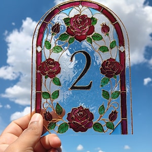 May include: An arched stained glass panel featuring a central black number 2 surrounded by a decorative border of deep red roses and green leaves. The glass has a textured surface and is framed by a dark red and gold metallic outline.