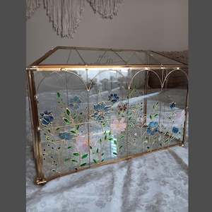May include: A rectangular glass display box with a gold metal frame featuring hand-painted floral designs in blue, pink, and green. The clear glass panels are decorated with delicate stems and blossoms, and the text The Adventures is visible.