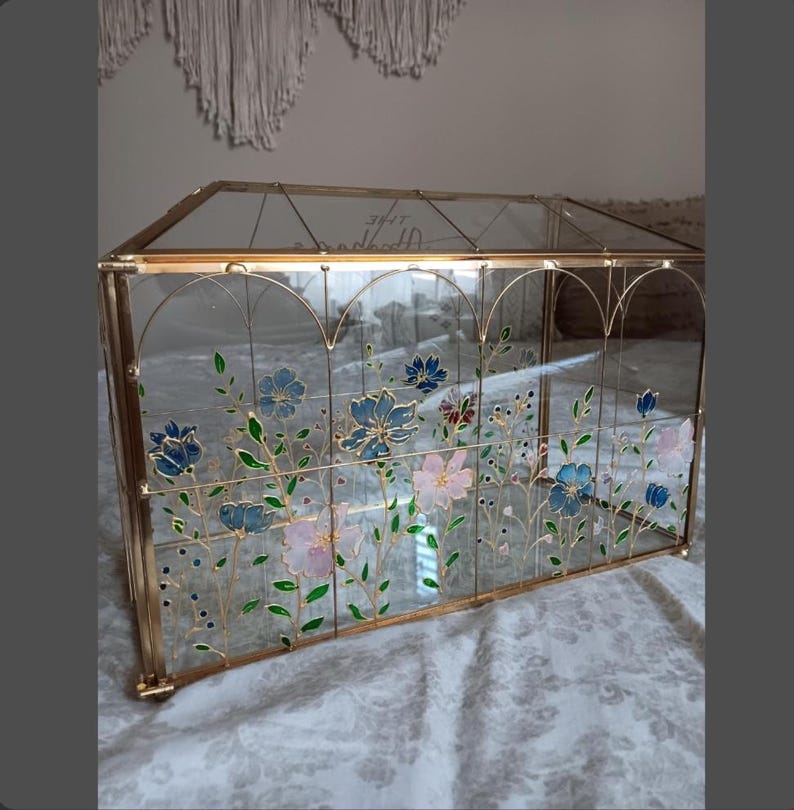May include: A rectangular glass display box with a gold metal frame featuring hand-painted floral designs in blue, pink, and green. The clear glass panels are decorated with delicate stems and blossoms, and the text The Adventures is visible.