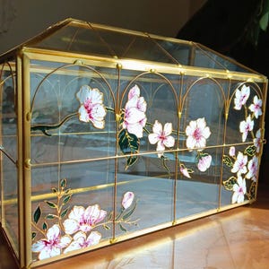 May include: A decorative glass terrarium with a gold-colored metal frame. The clear glass panels are adorned with hand-painted white and pink floral designs. The terrarium has a hinged door and a pitched roof, perfect for displaying plants or small items.