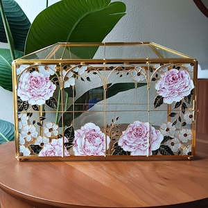 May include: A rectangular glass terrarium with a gold metal frame. The glass panels are decorated with pink peony flowers and white blossoms. The terrarium sits on a wooden surface.