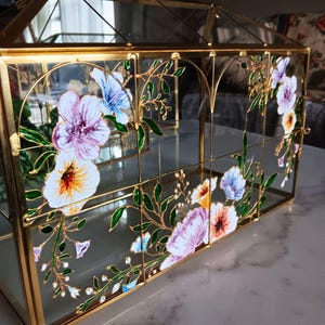 May include: A rectangular glass terrarium with a gold metal frame, featuring hand-painted floral designs in shades of purple, blue, white, and yellow with green leaves. The structure has a peaked roof and clear glass panels on all sides.