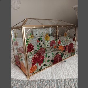 May include: A rectangular glass terrarium with a gold metal frame, featuring a pitched roof design. The clear glass panels are decorated with painted floral motifs in shades of red, orange, and yellow, accompanied by green leaves and stems.