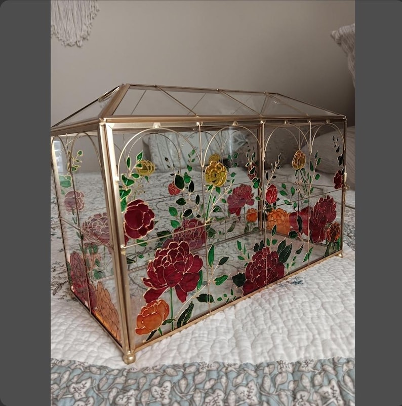 May include: A rectangular glass terrarium with a gold metal frame, featuring a pitched roof design. The clear glass panels are decorated with painted floral motifs in shades of red, orange, and yellow, accompanied by green leaves and stems.