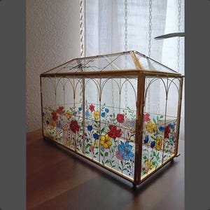 May include: A rectangular glass terrarium with a gold metal frame and a peaked roof. The glass panels are decorated with hand-painted colorful flowers in red, yellow, and blue. The text "The Miles" is written in black script on the top glass panel.