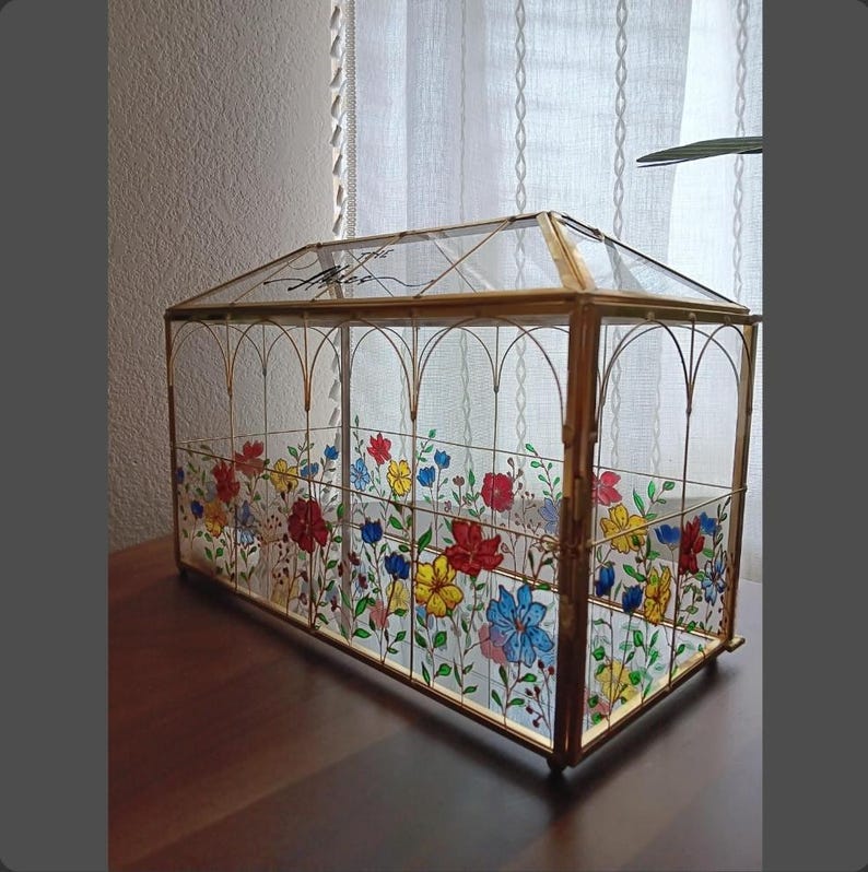 May include: A rectangular glass terrarium with a gold metal frame and a peaked roof. The glass panels are decorated with hand-painted colorful flowers in red, yellow, and blue. The text "The Miles" is written in black script on the top glass panel.