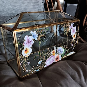 May include: A decorative glass terrarium with a gold metal frame. The glass panels are adorned with hand-painted floral designs in shades of purple, blue, yellow, and white. The terrarium has a hinged roof and is designed to house small plants.