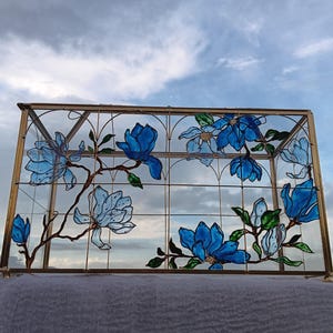 May include: A rectangular glass display box with a brass metal frame. The front glass panel features a hand-painted design of blue magnolia flowers with green leaves and brown branches, outlined in black, set against a clear glass background.