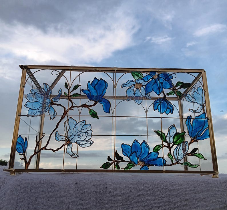 May include: A rectangular glass display box with a brass metal frame. The front glass panel features a hand-painted design of blue magnolia flowers with green leaves and brown branches, outlined in black, set against a clear glass background.