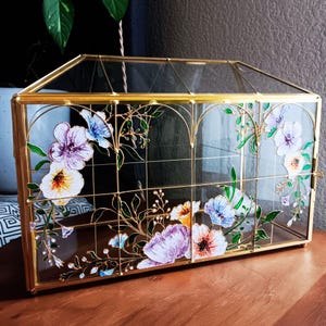 May include: A rectangular glass display box with a gold metal frame and a hinged lid. The glass panels are decorated with hand-painted floral designs in shades of purple, blue, orange, and green, featuring delicate stems and leaves.