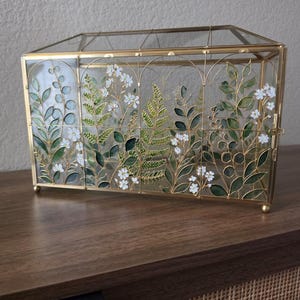 May include: A rectangular glass display box with a gold metal frame. The front glass panels feature a painted botanical design of green leaves, ferns, and small white flowers arranged within arched sections. The box has a hinged lid and small feet.