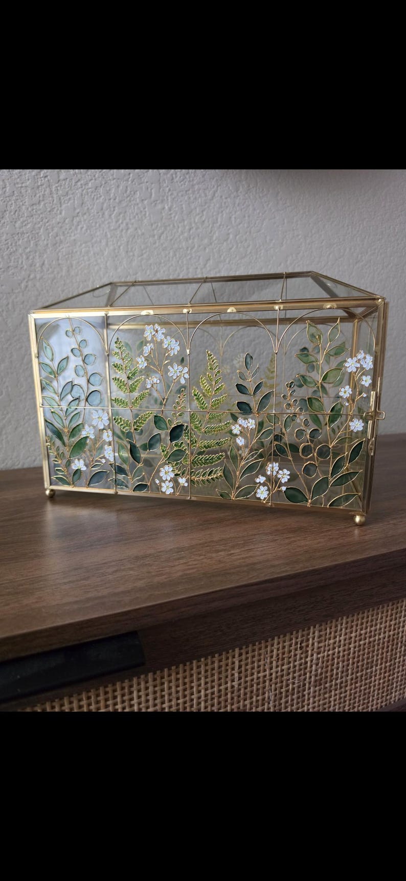 May include: A rectangular glass display box with a gold metal frame. The front glass panels feature a painted botanical design of green leaves, ferns, and small white flowers arranged within arched sections. The box has a hinged lid and small feet.