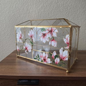May include: A rectangular glass display case with a gold metal frame, featuring hand-painted pink and white magnolia flowers with green leaves on the transparent panels. The structure has a peaked roof design and small gold ball feet.