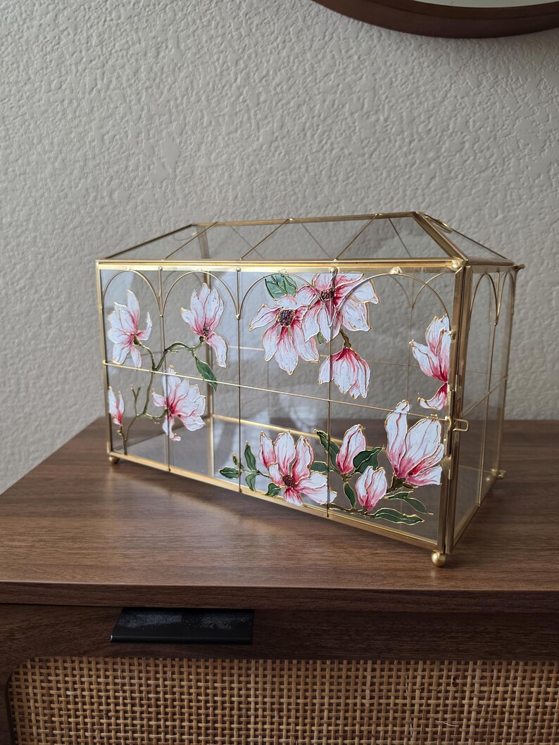 May include: A rectangular glass display case with a gold metal frame, featuring hand-painted pink and white magnolia flowers with green leaves on the transparent panels. The structure has a peaked roof design and small gold ball feet.