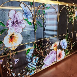 May include: A rectangular glass display case with a gold metal frame featuring arched top panels. The glass is decorated with hand-painted floral designs in shades of purple, white, and orange, accented by green leaves and thin gold outlines.