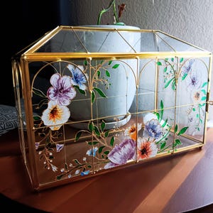 May include: A rectangular glass terrarium with a gold metal frame and a peaked roof. The clear glass panels are decorated with painted floral designs in shades of purple, blue, orange, and green, featuring delicate vines and various flower shapes.