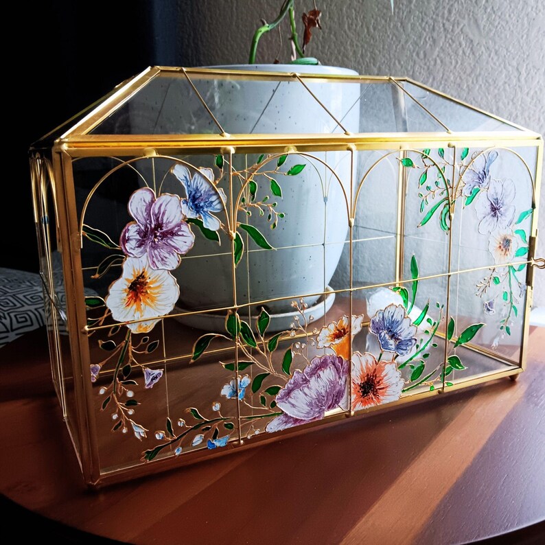 May include: A rectangular glass terrarium with a gold metal frame and a peaked roof. The clear glass panels are decorated with painted floral designs in shades of purple, blue, orange, and green, featuring delicate vines and various flower shapes.