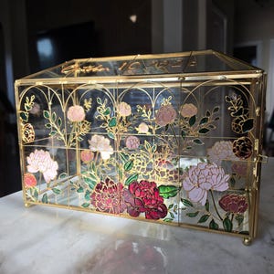 May include: A rectangular glass box with a gold metal frame, featuring hand-painted floral designs in shades of pink, red, and green. The top glass panel is etched with the text October 10 2024. The structure has a hinged lid and small feet.