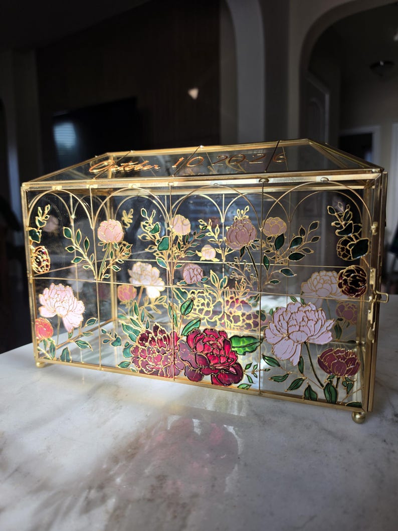 May include: A rectangular glass box with a gold metal frame, featuring hand-painted floral designs in shades of pink, red, and green. The top glass panel is etched with the text October 10 2024. The structure has a hinged lid and small feet.