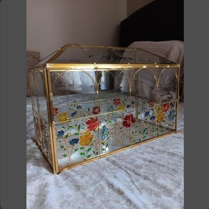 May include: A rectangular glass terrarium with a gold metal frame and a gabled roof. The clear glass panels are decorated with hand-painted colorful flowers in red, yellow, and blue with green stems and leaves. The word "Always" is written in black script on the top glass panel.