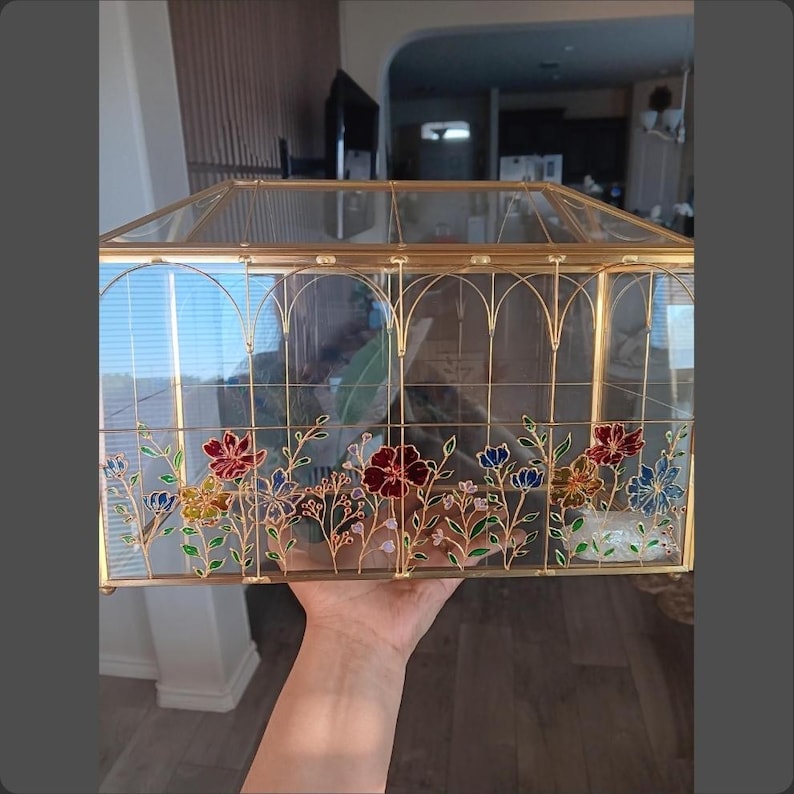 May include: A rectangular glass display box with a gold metal frame featuring arched top panels. The clear glass sides are decorated with painted floral designs in shades of red, blue, yellow, and green, depicting various stems and blossoms.