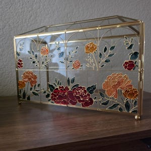 May include: A rectangular glass display box with a gold metal frame. The glass panels feature hand-painted floral designs in shades of deep red, orange, and yellow, accented with dark green leaves and thin gold outlines throughout the structure.